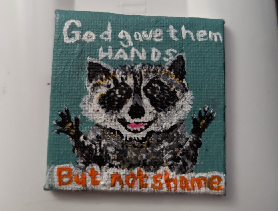 A 2 inch by 2 inch canvas on which is painted a little raccoon, mouth open so he looks like he’s smiling, with his little paws in the air. There is a teal background behind him. In white letters above his head, text reads God gave them hands. In Orange on white text below him, it says but not shame.
