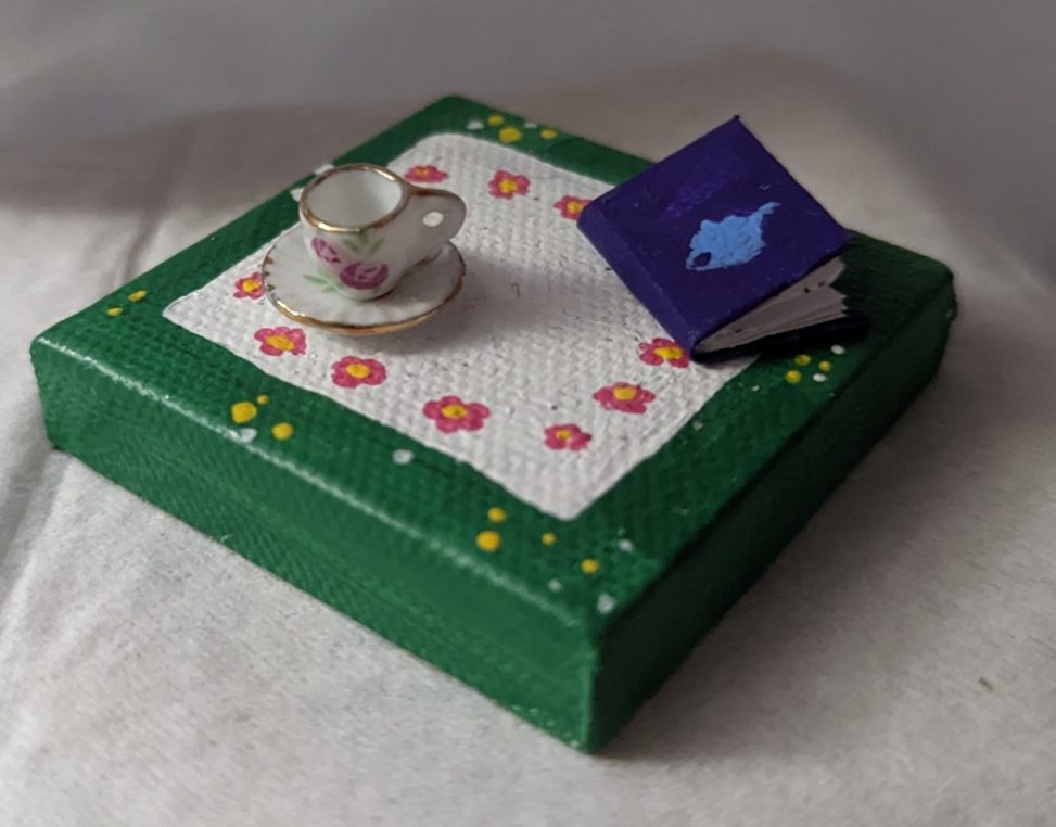Three images at various angles of a small canvas painted to look like a patch of grass with a small white cloth spread across it. There are pink flowers painted around the edges of the cloth. A three-dimensional teacup, also with a pink flower on it sits on the cloth, alongside a small purple book with a blue teapot painted on it.